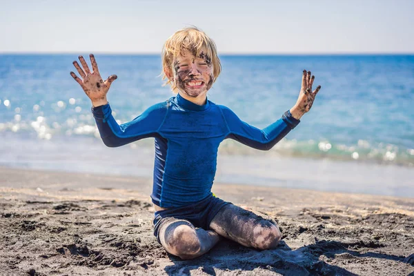 Concept Black Friday. Garçon souriant avec sale visage noir assis et jouant sur la plage de sable noir avant de nager dans l'océan. Mode de vie actif familial et loisirs aquatiques en vacances d'été avec les enfants — Photo