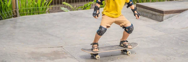 Atletik anak laki-laki dalam helm dan bantalan lutut belajar untuk skateboard dengan di sebuah taman skate. Pendidikan anak-anak, olahraga BANNER, LONG FORMAT — Stok Foto