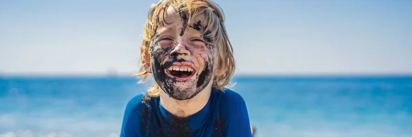 BANNER, FORMATO LUNGO concetto di Black Friday. Ragazzo sorridente con la faccia nera sporca seduto e giocare sulla spiaggia di sabbia nera prima di nuotare nell'oceano. Stile di vita attivo della famiglia e svago acquatico in estate — Foto Stock