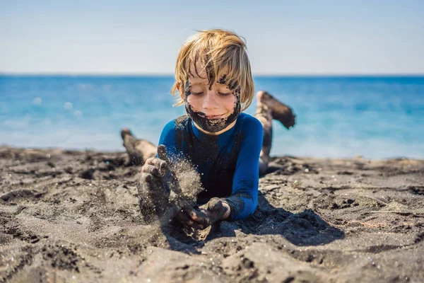 Koncepcja czarnego piątku. Uśmiechnięty chłopak z brudną czarną twarzą siedząc i grając na czarnym piasku morza plaży przed pływaniem w oceanie. Rodzinny aktywny styl życia i wodny wypoczynek na letnich wakacjach z dziećmi — Zdjęcie stockowe