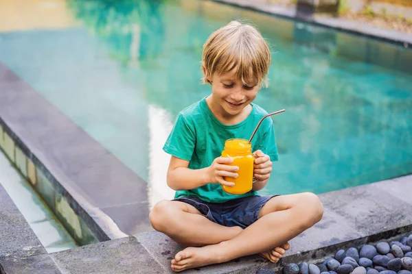 Boy drinking juicy smoothie from mango in glass mason jar with steel straw on the background of the pool. Healthy life concept, copy space