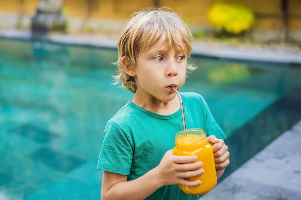 Pojke dricker saftig smoothie från mango i glas Mason burk med stål halm på bakgrunden av poolen. Hälsosamt liv koncept, kopiera utrymme — Stockfoto