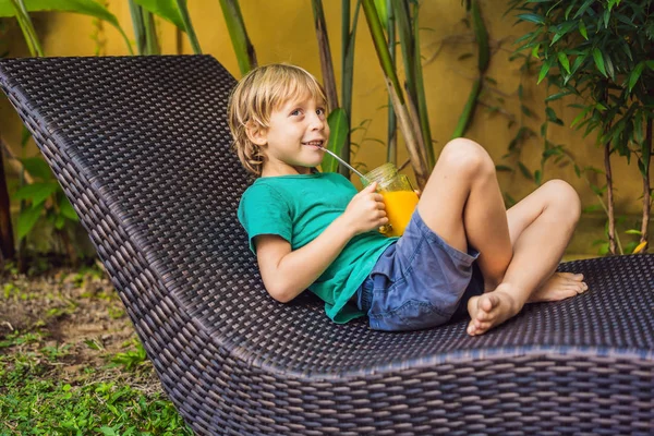 Boy drinking juicy smoothie from mango in glass mason jar with steel straw on the background of the pool. Healthy life concept, copy space