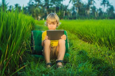 Sahada tablet tutan mutlu çocuk. Güneşli bir günde çimenlerin üzerinde oturan çocuk. Evde eğitim veya tablet oynama