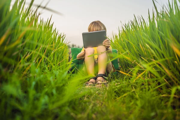 Joyeux enfant assis sur le terrain tenant la tablette. Garçon assis sur l'herbe par une journée ensoleillée. Enseignement à domicile ou lecture d'une tablette — Photo