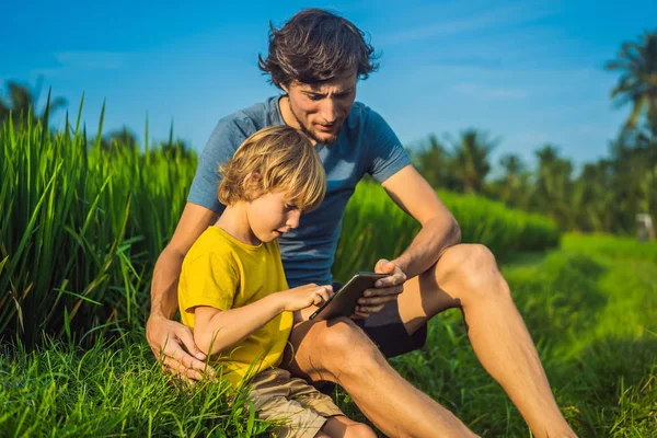 Dad and son sitting on the field holding tablet. Boy sitting on the grass on sunny day. Home schooling or playing a tablet — Stock Photo, Image