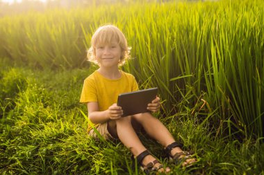 Sahada tablet tutan mutlu çocuk. Güneşli bir günde çimenlerin üzerinde oturan çocuk. Evde eğitim veya tablet oynama