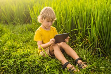 Sahada tablet tutan mutlu çocuk. Güneşli bir günde çimenlerin üzerinde oturan çocuk. Evde eğitim veya tablet oynama