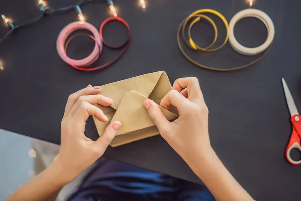 A jovem está a empacotar presentes. Presente embrulhado em papel artesanal com uma fita vermelha e dourada para o Natal, aniversário, dia das mães ou dia dos namorados — Fotografia de Stock