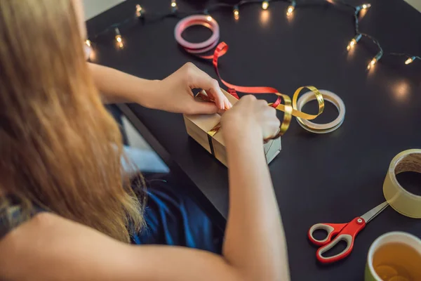 A jovem está a empacotar presentes. Presente embrulhado em papel artesanal com uma fita vermelha e dourada para o Natal, aniversário, dia das mães ou dia dos namorados — Fotografia de Stock