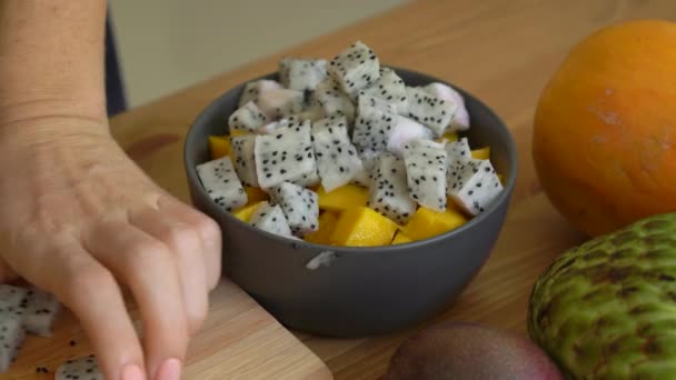 Una joven mujer poniendo los cubos de fruta de dragón en un tazón de cerámica gris y un montón de frutas tropicales yacen en una mesa — Vídeo de stock