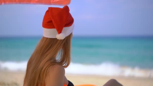 Super Slow Motion shot van een jonge vrouw in een kerstmuts ontspannen onder een paraplu op een opblaasbare bank op een tropisch strand. — Stockvideo