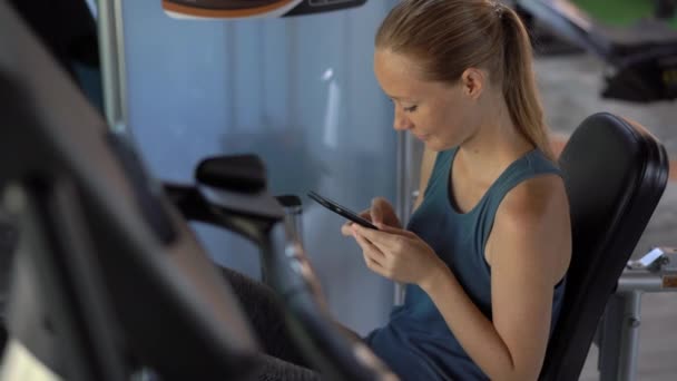 Uma jovem mulher no ginásio fazendo exercícios segurando um telefone na frente de seus olhos em suas mãos. Toda a atenção está no telefone. O conceito de dependência das redes sociais. Conceito de dependência móvel — Vídeo de Stock