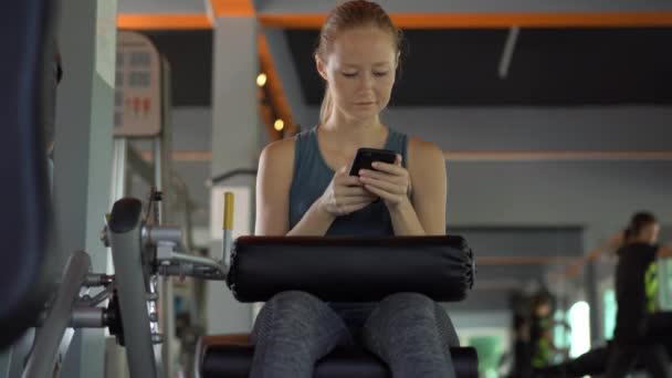 Μια νεαρή γυναίκα στο γυμναστήριο κάνει ασκήσεις κρατώντας ένα τηλέφωνο μπροστά στα μάτια της στα χέρια της. Όλη η προσοχή είναι στο τηλέφωνο. Η έννοια της εξάρτησης από τα κοινωνικά δίκτυα. Ιδέα για το κινητό εθισμό — Αρχείο Βίντεο