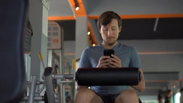 Un joven en el gimnasio haciendo ejercicios sosteniendo un teléfono delante de sus ojos en sus manos. Toda la atención está en el teléfono. El concepto de dependencia de las redes sociales. Concepto de adicción móvil — Vídeos de Stock