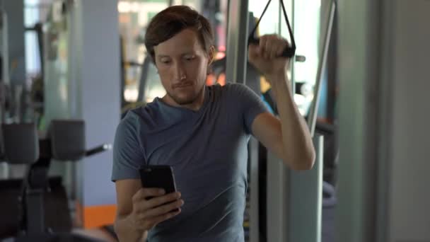 Un joven en el gimnasio haciendo ejercicios sosteniendo un teléfono delante de sus ojos en sus manos. Toda la atención está en el teléfono. El concepto de dependencia de las redes sociales. Concepto de adicción móvil — Vídeo de stock