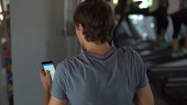 Un joven en el gimnasio haciendo ejercicios sosteniendo un teléfono delante de sus ojos en sus manos. Toda la atención está en el teléfono. El concepto de dependencia de las redes sociales. Concepto de adicción móvil — Vídeo de stock