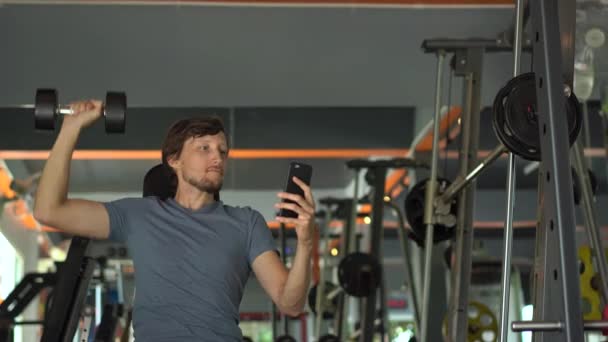 Ένας νεαρός στο γυμναστήριο που κάνει ασκήσεις κρατώντας ένα τηλέφωνο μπροστά στα μάτια του στα χέρια του. Όλη η προσοχή είναι στο τηλέφωνο. Η έννοια της εξάρτησης από τα κοινωνικά δίκτυα. Ιδέα για το κινητό εθισμό — Αρχείο Βίντεο