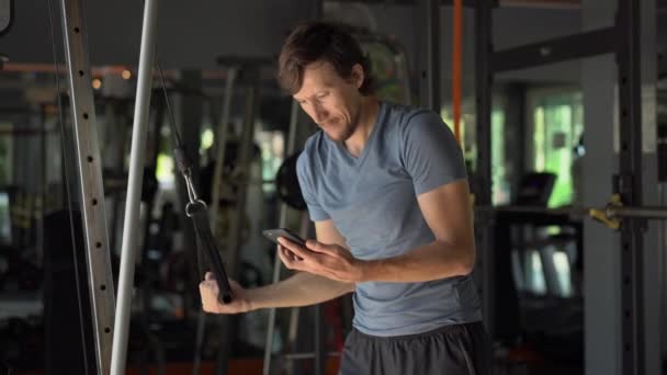 Een jonge man in de sportschool doen oefeningen houden een telefoon voor zijn ogen in zijn handen. Alle aandacht is in de telefoon. Het begrip afhankelijkheid van sociale netwerken. Mobiel verslavings concept — Stockvideo