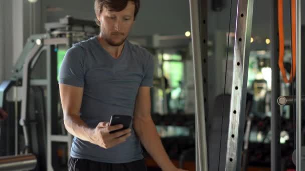 Een jonge man in de sportschool doen oefeningen houden een telefoon voor zijn ogen in zijn handen. Alle aandacht is in de telefoon. Het begrip afhankelijkheid van sociale netwerken. Mobiel verslavings concept — Stockvideo