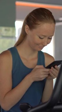 Dikey video. Spor salonunda genç bir kadın elinde gözlerinin önünde bir telefon tutarak egzersiz yapıyor. Tüm dikkatler telefonda. Sosyal ağlara bağımlılık kavramı. Mobil