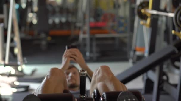 Un joven en el gimnasio haciendo ejercicios sosteniendo un teléfono delante de sus ojos en sus manos. Toda la atención está en el teléfono. El concepto de dependencia de las redes sociales. Concepto de adicción móvil — Vídeos de Stock