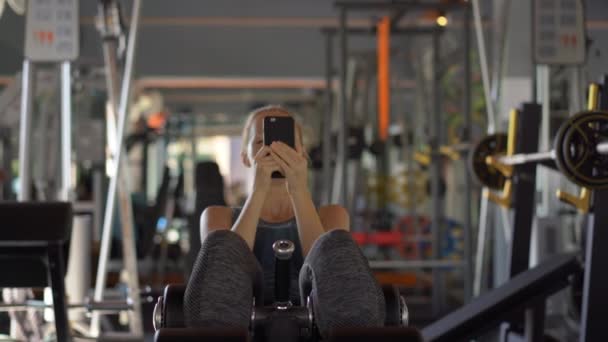 Μια νεαρή γυναίκα στο γυμναστήριο κουνάει τον τύπο κρατώντας το τηλέφωνο μπροστά στα μάτια της στα χέρια της. Όλη η προσοχή είναι στο τηλέφωνο. Η έννοια της εξάρτησης από τα κοινωνικά δίκτυα. Ιδέα για το κινητό εθισμό — Αρχείο Βίντεο