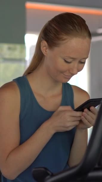 Vídeo vertical. Uma jovem mulher no ginásio fazendo exercícios segurando um telefone na frente de seus olhos em suas mãos. Toda a atenção está no telefone. O conceito de dependência das redes sociais. Móvel — Vídeo de Stock