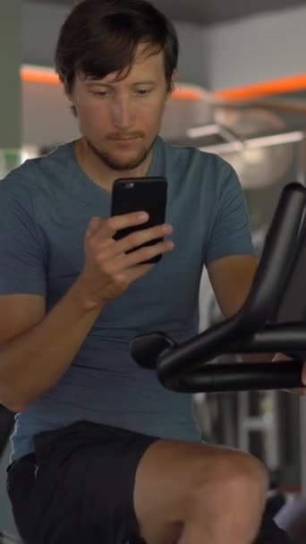 Vídeo vertical. Un joven en el gimnasio en una bicicleta estática sosteniendo el teléfono frente a sus ojos en sus manos. Toda la atención está en el teléfono. El concepto de dependencia de las redes sociales. Móvil — Vídeo de stock