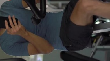 Dikey video. Spor salonunda bir egzersiz bisikleti üzerinde genç bir adam elinde gözlerinin önünde telefon tutarak. Tüm dikkatler telefonda. Sosyal ağlara bağımlılık kavramı. Mobil