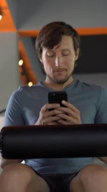 Dikey video. Spor salonunda bir egzersiz bisikleti üzerinde genç bir adam elinde gözlerinin önünde telefon tutarak. Tüm dikkatler telefonda. Sosyal ağlara bağımlılık kavramı. Mobil