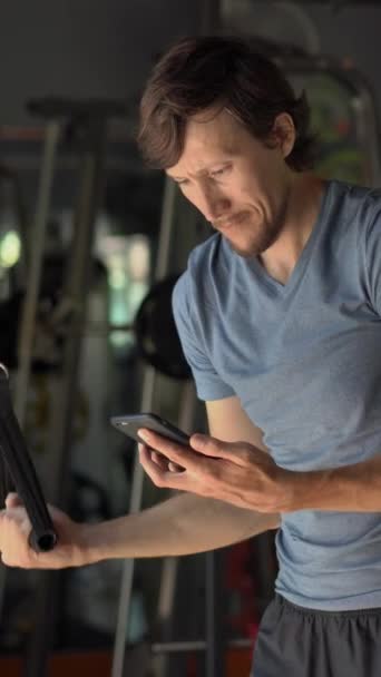 Wideo w pionie. Młody człowiek na siłowni na rowerze treningowym trzymając telefon przed oczami w dłoniach. Cała Uwaga jest w telefonie. Pojęcie uzależnienia od sieci społecznościowych. Mobile — Wideo stockowe