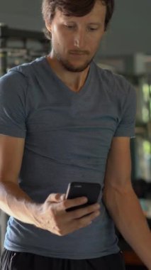 Dikey video. Spor salonunda bir egzersiz bisikleti üzerinde genç bir adam elinde gözlerinin önünde telefon tutarak. Tüm dikkatler telefonda. Sosyal ağlara bağımlılık kavramı. Mobil