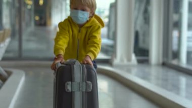 Little boy in a medical face mask playing with a suitcase in an airport