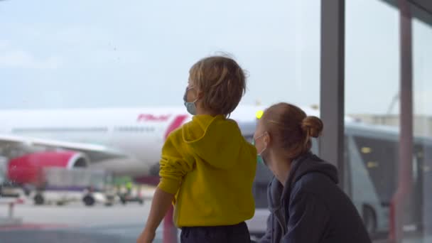 Kleiner Junge und seine Mutter mit medizinischer Gesichtsmaske auf einem Flughafen, der auf ihren Flug wartet — Stockvideo