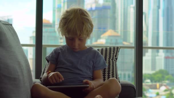 Niño pequeño usando una tableta PC sentado frente a una gran ventana con rascacielos en un fondo — Vídeo de stock