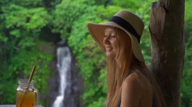 Bir kanyon ve şelale üzerinde destansı bir görünümü ile güzel bir kafede genç bir kadının slowmotion çekim buz ile çay içecekler. Travell bali kavramı