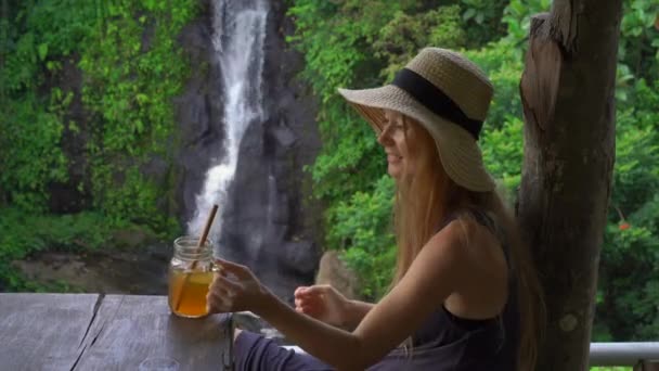 Uma jovem em um belo café com uma vista épica sobre um desfiladeiro e cachoeira bebe chá com gelo. Conceito de Travell para Bali — Vídeo de Stock