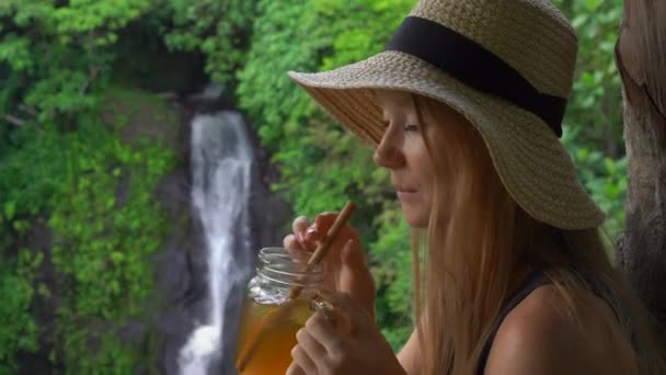 Uma jovem em um belo café com uma vista épica sobre um desfiladeiro e cachoeira bebe chá com gelo. Conceito de Travell para Bali — Vídeo de Stock