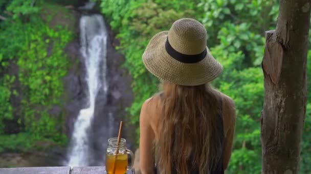 Una joven en un hermoso café con una vista épica de un cañón y una cascada bebe té con hielo. Viajar al concepto de Bali — Vídeo de stock