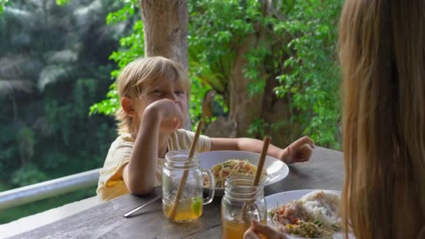 Η νεαρή γυναίκα και ο μικρός της γιος έχουν ένα Λαντς σε ένα καφέ με επική θέα σε ένα φαράγγι και καταρράκτη. Ταξίδι στο Μπαλί — Αρχείο Βίντεο