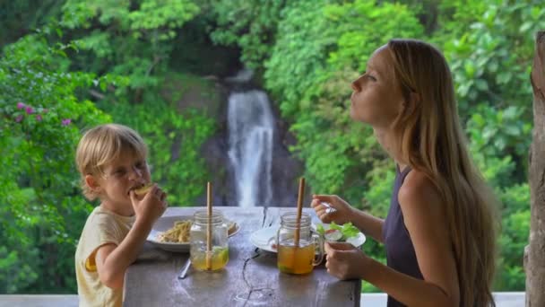Молодая женщина и ее маленький сын имеют ланч в кафе с эпическим видом на каньон и водопад. Концепция путешествия на Бали — стоковое видео