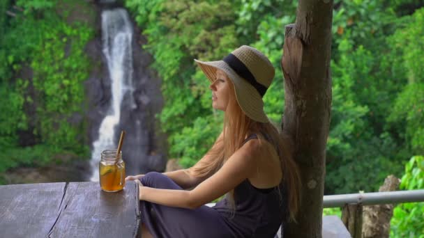 Slowmotion tiro de uma jovem mulher em um belo café com uma vista épica sobre um canyon e cachoeira bebe chá com gelo. Conceito de Travell para Bali — Vídeo de Stock