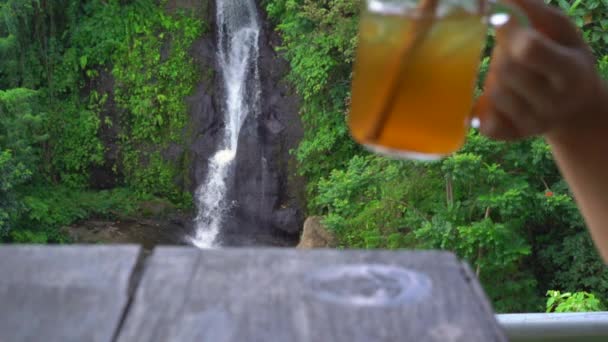 Foto en cámara lenta de una mujer joven en un hermoso café con una vista épica de un cañón y cascada bebe té con hielo. Viajar al concepto de Bali — Vídeos de Stock