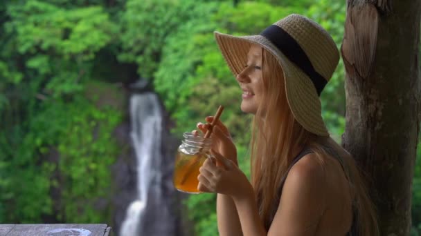 Slowmotion shot van een jonge vrouw in een prachtig café met een episch uitzicht op een kloof en waterval drinkt thee met ijs. Travell naar Bali concept — Stockvideo