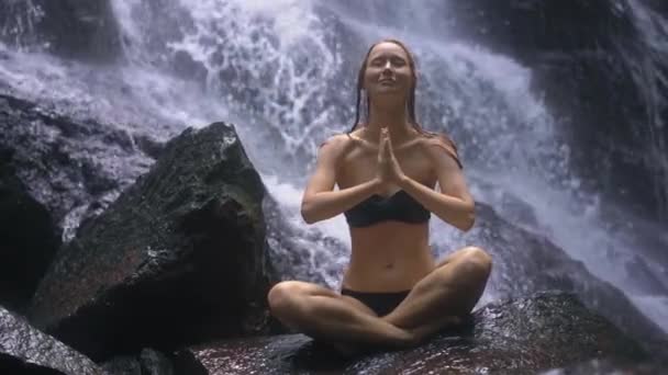 Junge Frau meditiert auf einem Felsen sitzend, hinter dem ein Wasserfall plätschert — Stockvideo