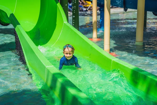 Niño feliz en el tobogán acuático en una piscina que se divierte durante las vacaciones de verano en un hermoso complejo tropical —  Fotos de Stock