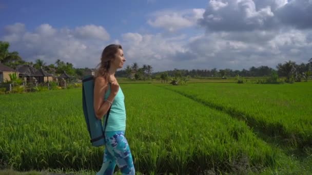 Slowmotion Steadicam shot van een jonge vrouw met yoga math wandelen door een prachtige rijstveld — Stockvideo