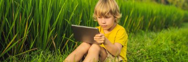 Sahada tablet tutan mutlu çocuk. Güneşli bir günde çimenlerin üzerinde oturan çocuk. Evde eğitim veya tablet Banner, Uzun Format oynama