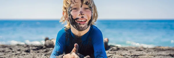 BANNER, LONGO FORMAT Conceito Black Friday. Garoto sorridente com rosto preto sujo sentado e brincando na praia de areia preta antes de nadar no oceano. Estilo de vida familiar ativo e lazer aquático no verão — Fotografia de Stock